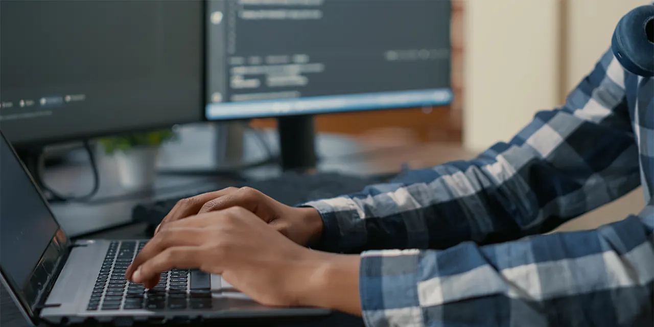Mãos digitando em um notebook com código aberto no monitor ao lado